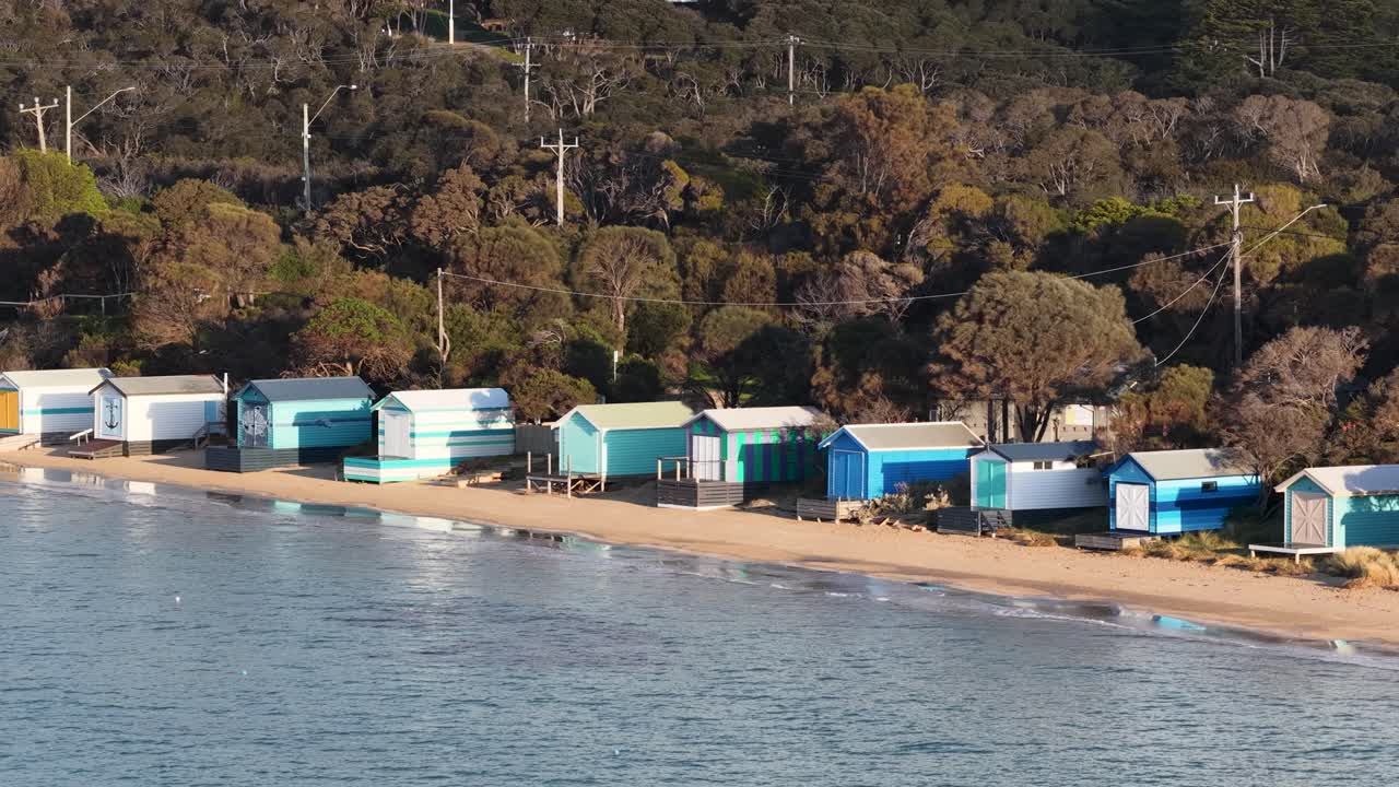 Drone pans over vibrant beach huts on sandy shore, morning sunlight, tranquil coastal environment