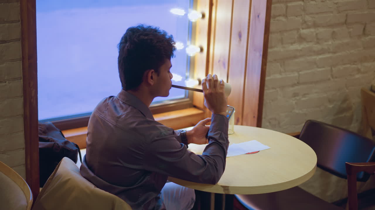 Young man sitting in cafe near window with mirror lights, holding empty paper cup while using smartphone, sipping last drop through straw, relaxed posture, calm indoor ambiance, city view outside