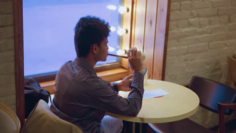 young man sitting in cafe near window with mirror lights, holding empty paper cup while using smartphone, sipping last drop through straw, relaxed posture, calm indoor ambiance, city view outside