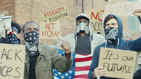 afroamerikanischer mann in gasmaske, der die flagge der vereinigten staaten aus protest gegen rassismus mit multiethnischer gruppe von menschen auf der straße hält