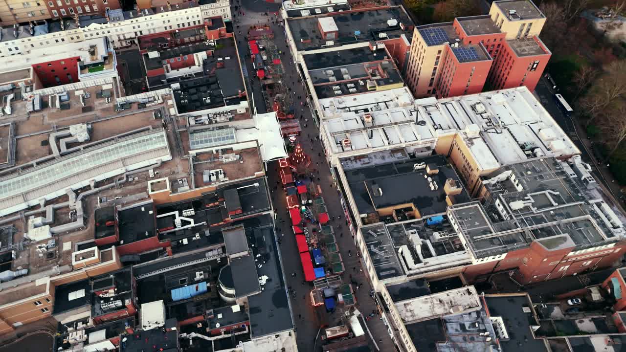 Aerial drone shot panning left over Southampton’s Christmas Market in winter at sunset, showcasing glowing festive stalls, bustling crowds, and the surrounding city bathed in warm evening light