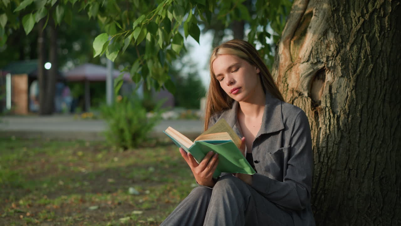 dama con ropa gris sentada al aire libre hojeando un libro con la cabeza inclinada ligeramente a la derecha, mirando pensativa, el fondo presenta vegetación y edificio borroso