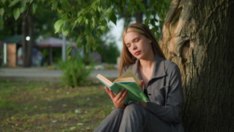 lady in grey clothing seated outdoors flipping through a book with her head tilted slightly to the right, looking thoughtful, background features greenery and blurred building