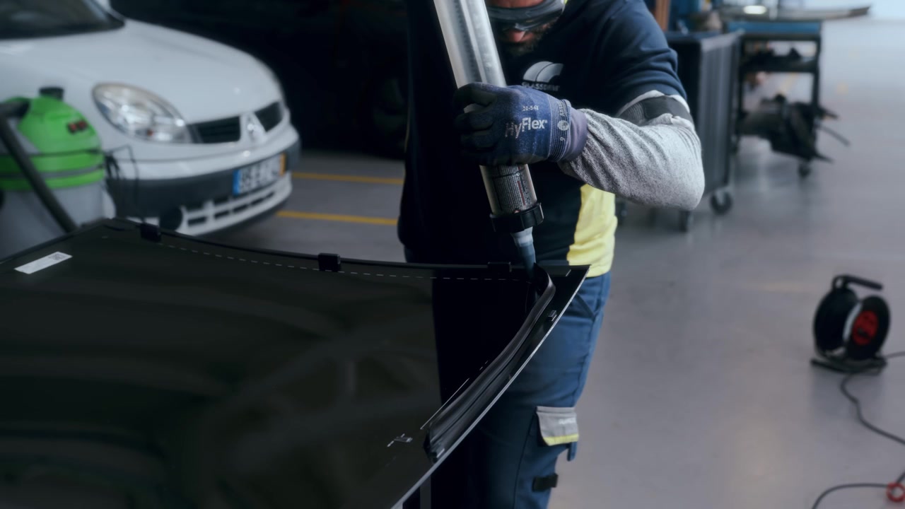 Handskeklædt arbejder påfører fugemasse med fugepistol langs kanten af en bilforrude.