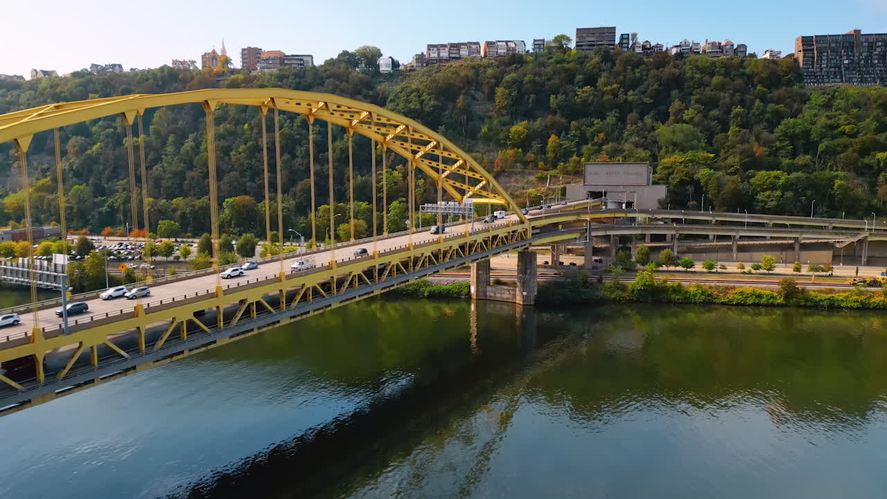 Transport moves to the Fort Duquesne Bridge in Pittsburgh, Pennsylvania, USA. Green hilly waterfront with city skyline on top at backdrop. Drone footage over the Allegheny River