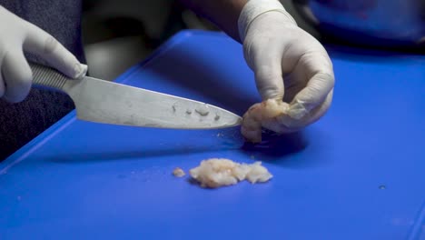 chef in black uniform, wearing white sterilized gloves is skillfully peeling, cleaning, deveining and cutting shrimp with a sharp knife on a blue plastic cutting board