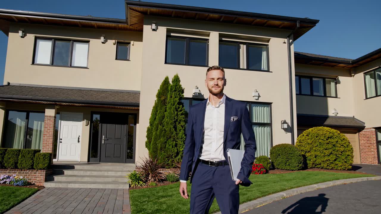 A confident man in a suit stands in front of a modern house, captured from a low-angle shot