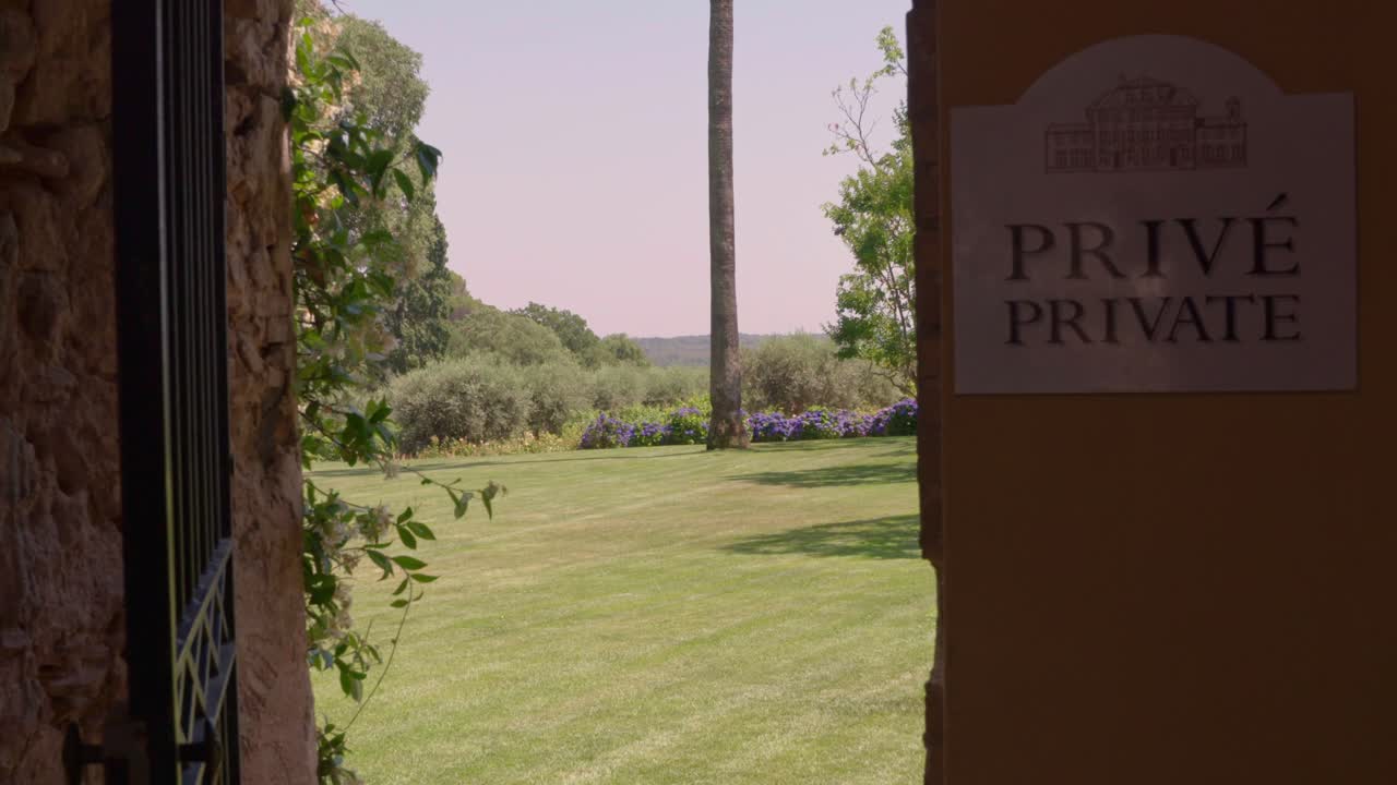 A wide shot captures an iron gate opening to reveal a serene courtyard in Italy, featuring a lush lawn, tall trees, and vibrant purple flowers under a clear sky.