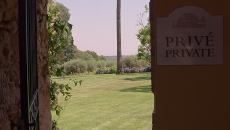 a wide shot captures an iron gate opening to reveal a serene courtyard in italy, featuring a lush lawn, tall trees, and vibrant purple flowers under a clear sky.