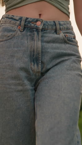 close-up of woman wearing high-waisted jeans and crop top, revealing her stomach, walking outdoors with blurred background of greenery, power lines, and urban structure