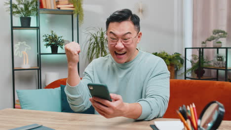 asian chinese man at home use smartphone celebrating success victory winning play online game