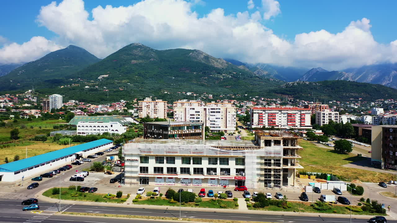 Flying above the blocks of flats and highway in the city of Bar, Montenegro. Green mountains with white cloudscape on top at backdrop. Aerial view.
