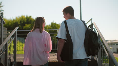 back view of friends walking up outdoor staircase in bright sunlight, interacting warmly, carrying backpacks, with greenery and modern structures in the background