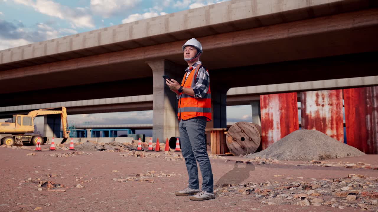 Full Body Side View Of Asian Male Engineer With Safety Helmet Using Smartphone And Looking Around While Standing At The Site of Highway Construction