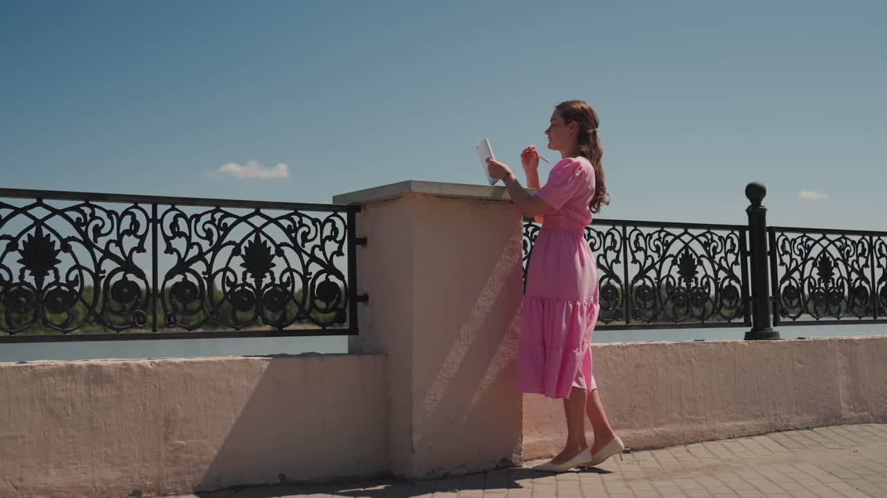 Mulher caucasiana de vestido rosa à beira-mar olhando através de uma grade de ferro ornamentada para um rio calmo, cabelo trançado, céu iluminado pelo sol, calçadão tranquilo e brisa suave criando um clima romântico cinematográfico para viagem