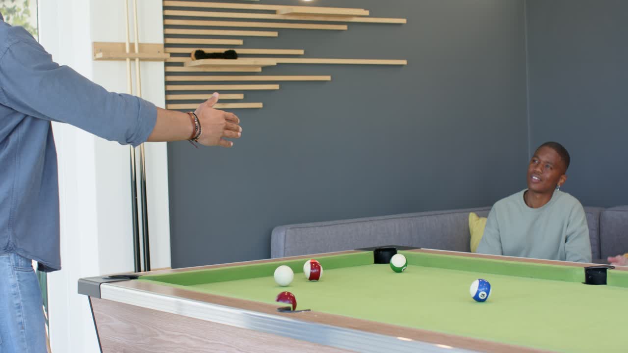Man approaching pool table and striking ball with cue stick then shaking friend's hand in lounge