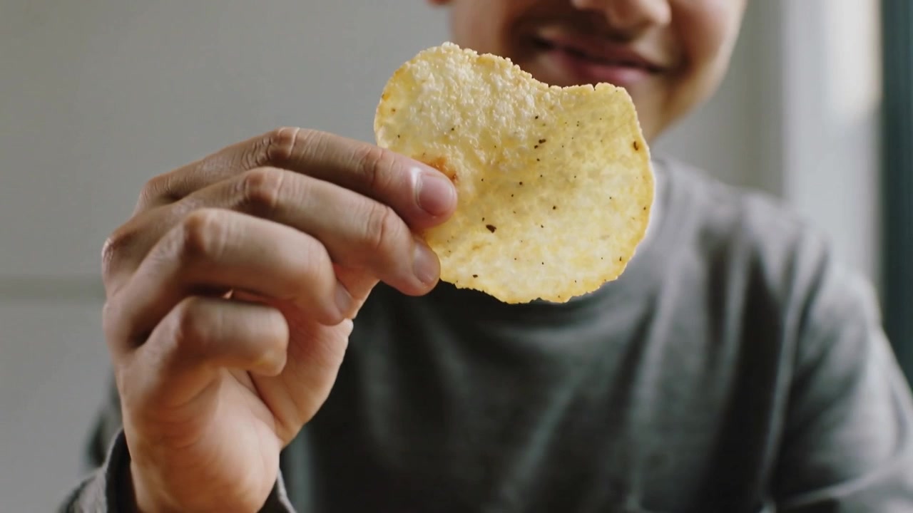 Reaching into bright chip bag, teen-male in gray shirt holding ridged chip on wood table, showing