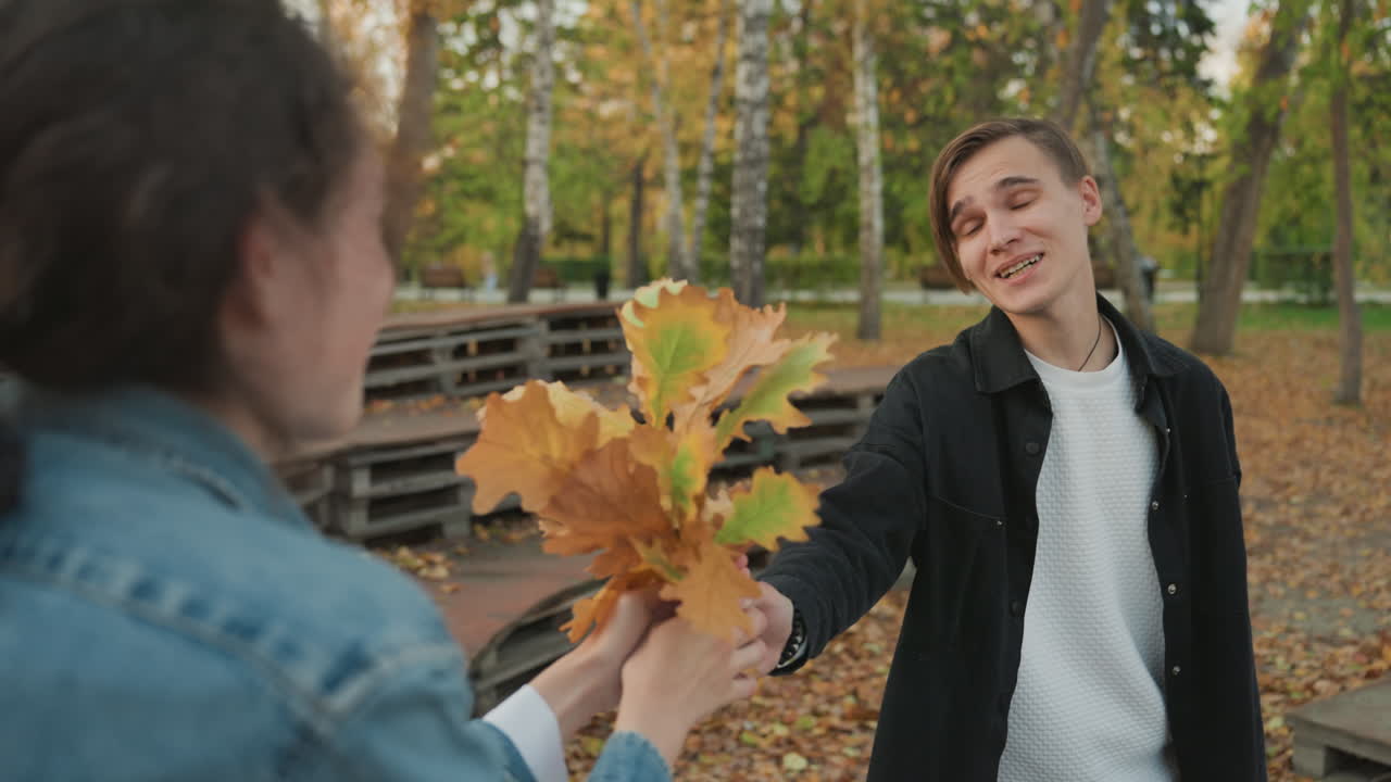 Ungt hvidt par udveksler efterårsblade på parkbænke, legende kurtisescene med smilende ung mand, der modtager en buket gyldne blade, hyggeligt gyldent aftenlys, afslappet denim og sweater