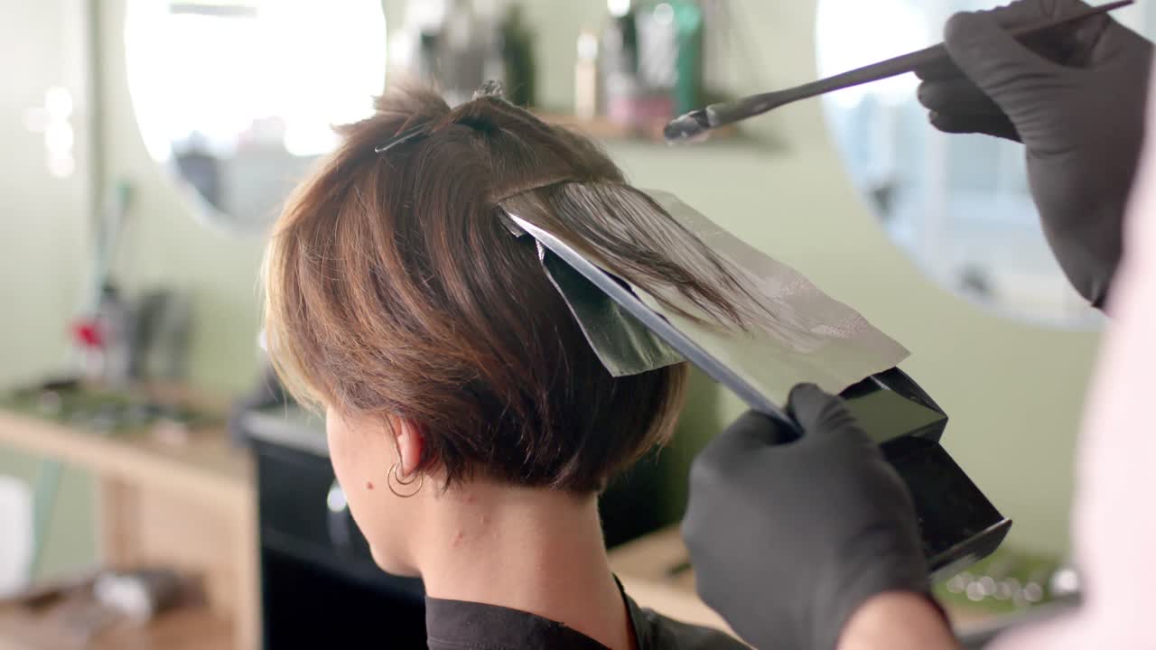 Hands of hairdresser highlighting client's hair with brush and foil at hair salon, in slow motion