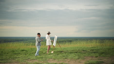 a woman in a white dress holds a palette and paints on a canvas while an artist in a black hat and checkered shirt stands beside her, holding a brush, in a peaceful grass field under a cloudy sky