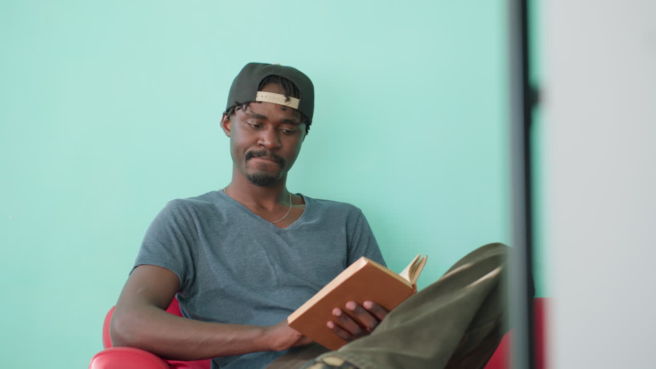 Student in casual shirt and cap sits on red couch with crossed leg, holding open book while reading calmly in bright indoor space with turquoise wall, focused on page