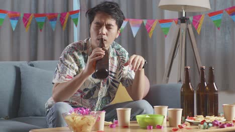 hombre asiático borracho sentado en un sofá y bebiendo alcohol mientras tiene una fiesta solo en casa