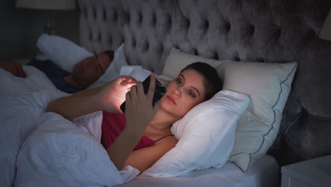 mujer acostada en la cama comprobando el teléfono móvil mientras el hombre duerme a su lado