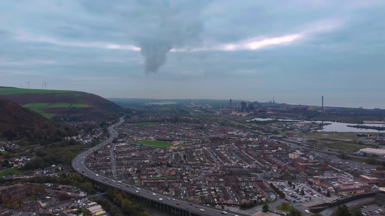 Ampia veduta aerea del paesaggio della città gallese di Port Talbot con nuvole scure sopra, con l'autostrada M4 che si snoda attraverso case e abitazioni suburbane e la fabbrica siderurgica sullo sfondo. 4K