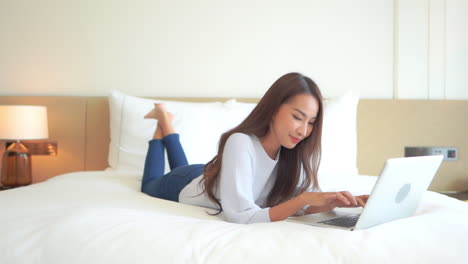 Young-Happy-Asian-Woman-Laying-on-Bed-and-Typing-on-Laptop