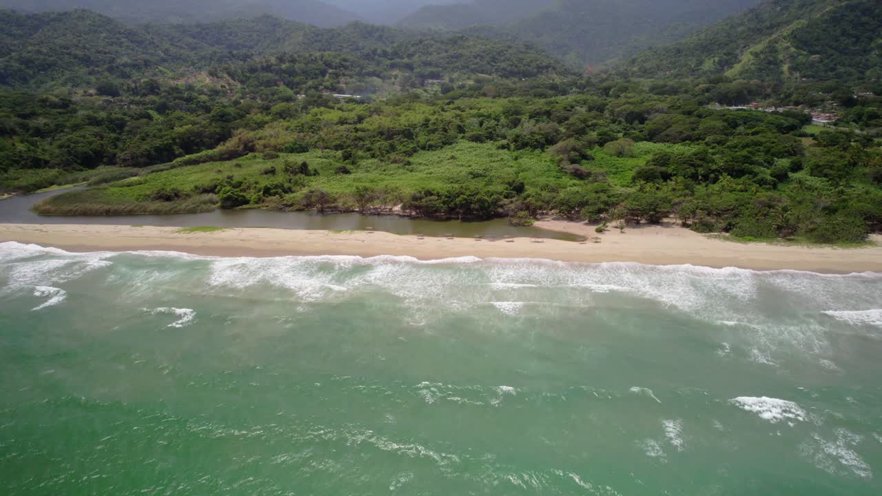 An empty tropical beach with waves crashing and green mountains in the background, aerial view