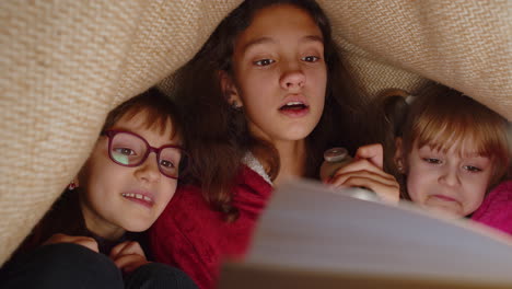 children sisters girls under plaid reading interesting fairytale story book using flashlight at home