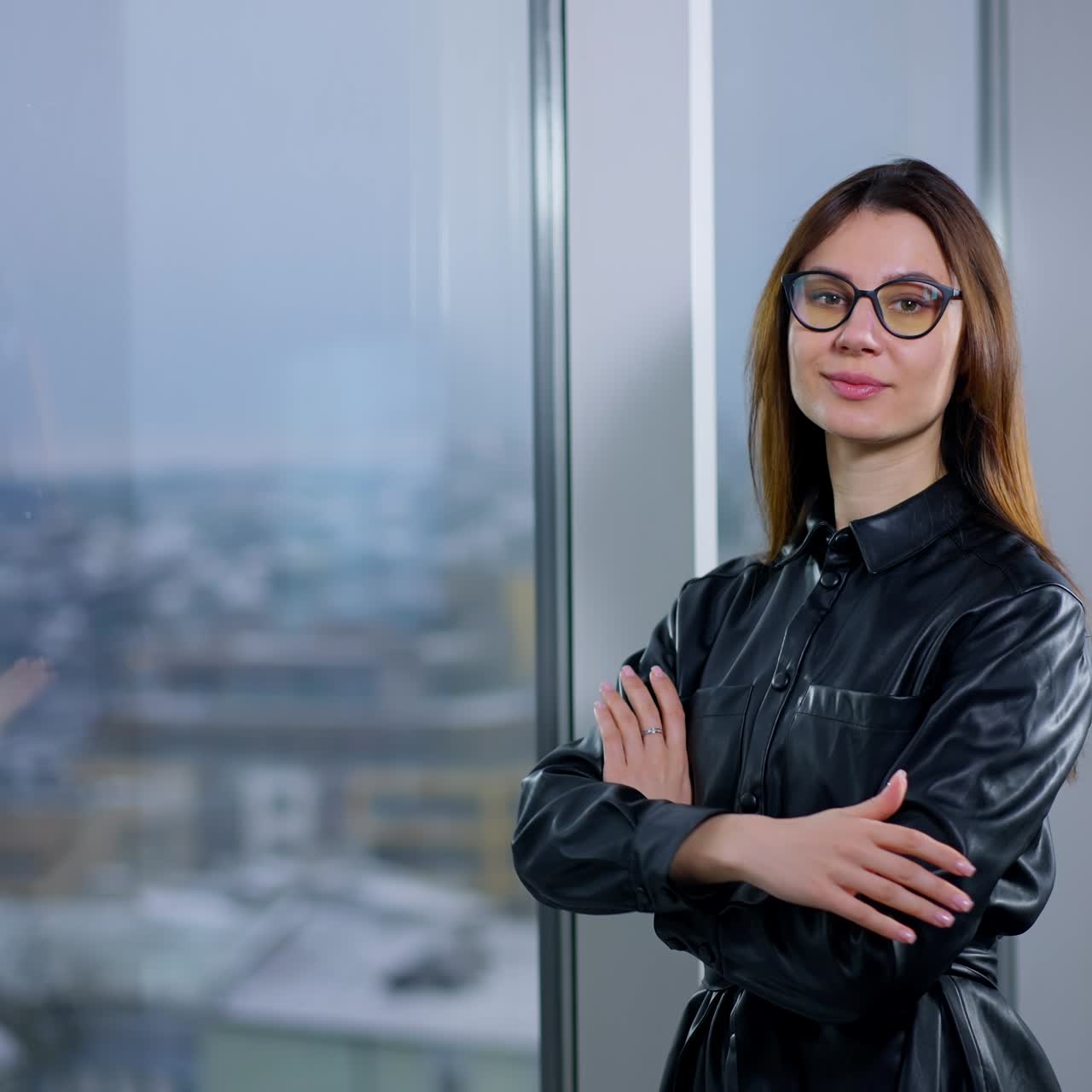 Long-haired brunette lady stands at the window. Woman puts on glasses, folding hands on her chest and looks at camera. Grey landscape in blur at backdrop