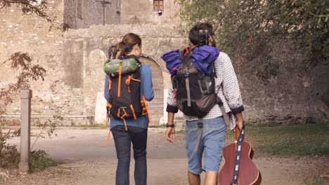 happy young couple backpackers tourists walking toward medieval building with wooden doorway in parco degli acquedotti park ruins in rome at sunrise with guitar and sleeping bag slow motion steadycam