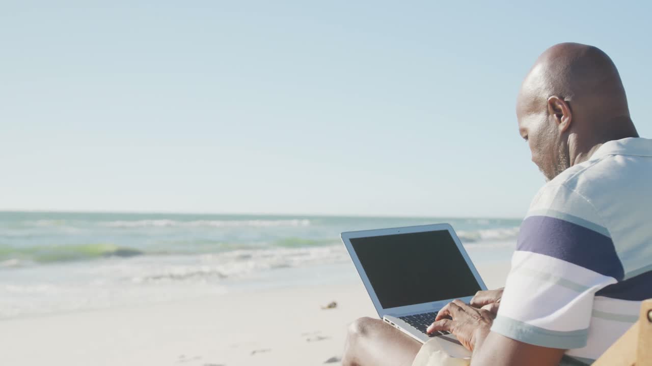 Senior african american man on deck chair and using laptop at beach, copy space, in slow motion