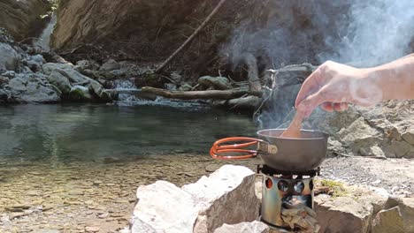 chico cocinero en la naturaleza, revolviendo la sopa en la cocina de campamento vagabundo con ríos y cascadas en el fondo