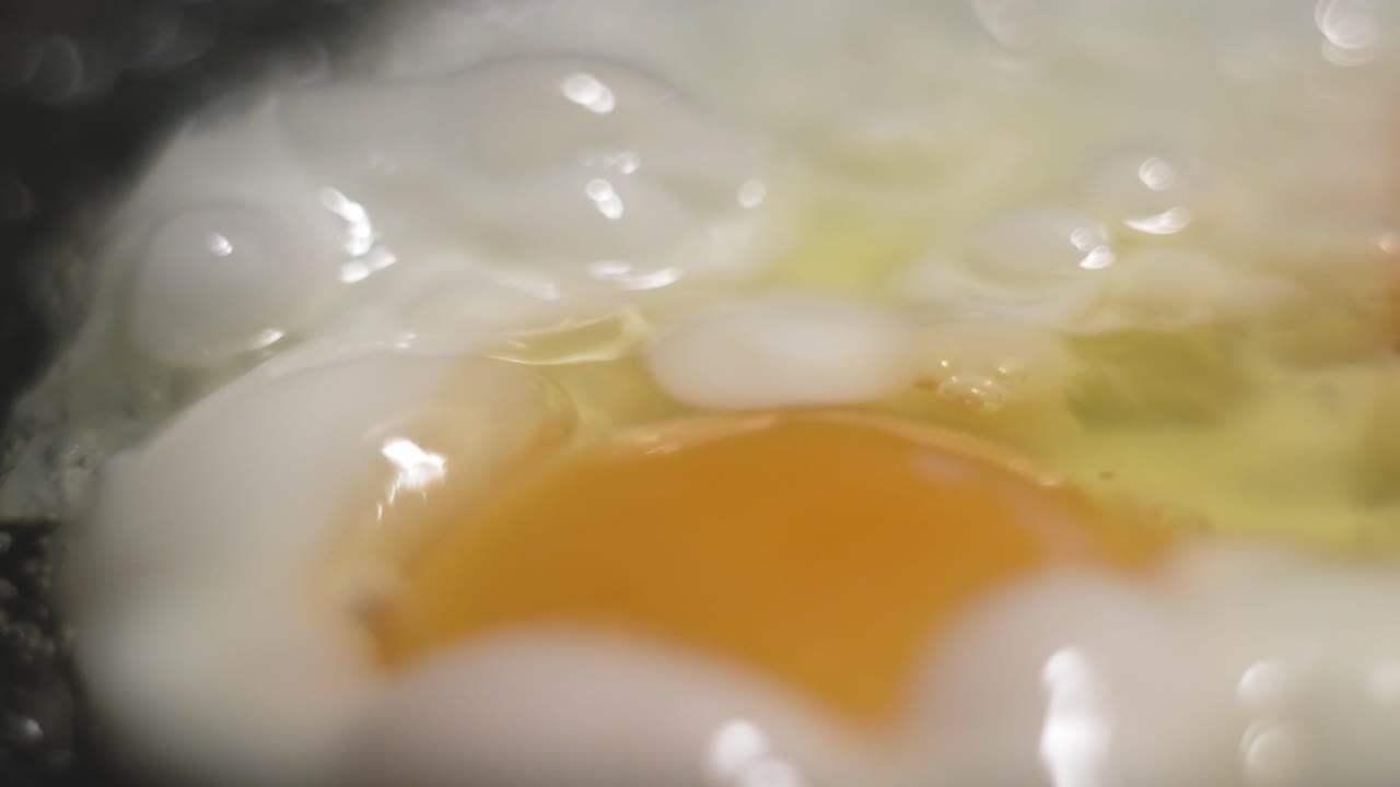 Frying eggs for breakfast in a black iron pan with bubbles jumping in hot oil as a soft background with bokeh