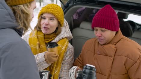 fröhliche freunde, die während einer winterreise in der nähe des autos in einem verschneiten wald zusammen trinken und reden