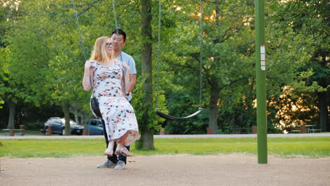 active young family resting in the park ride on a swing pregnant woman and asian man