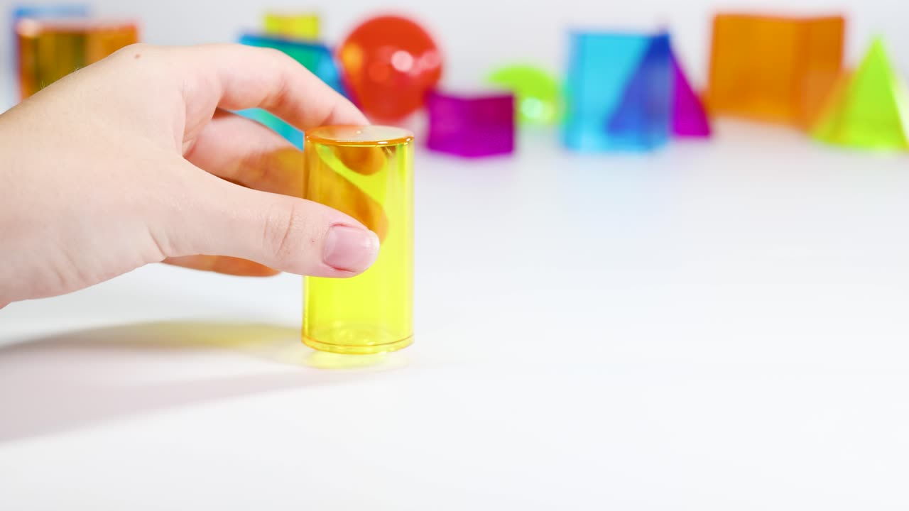 A hand manipulates vibrant 3D shapes on a white surface, highlighting geometry and color contrast in a playful setting
