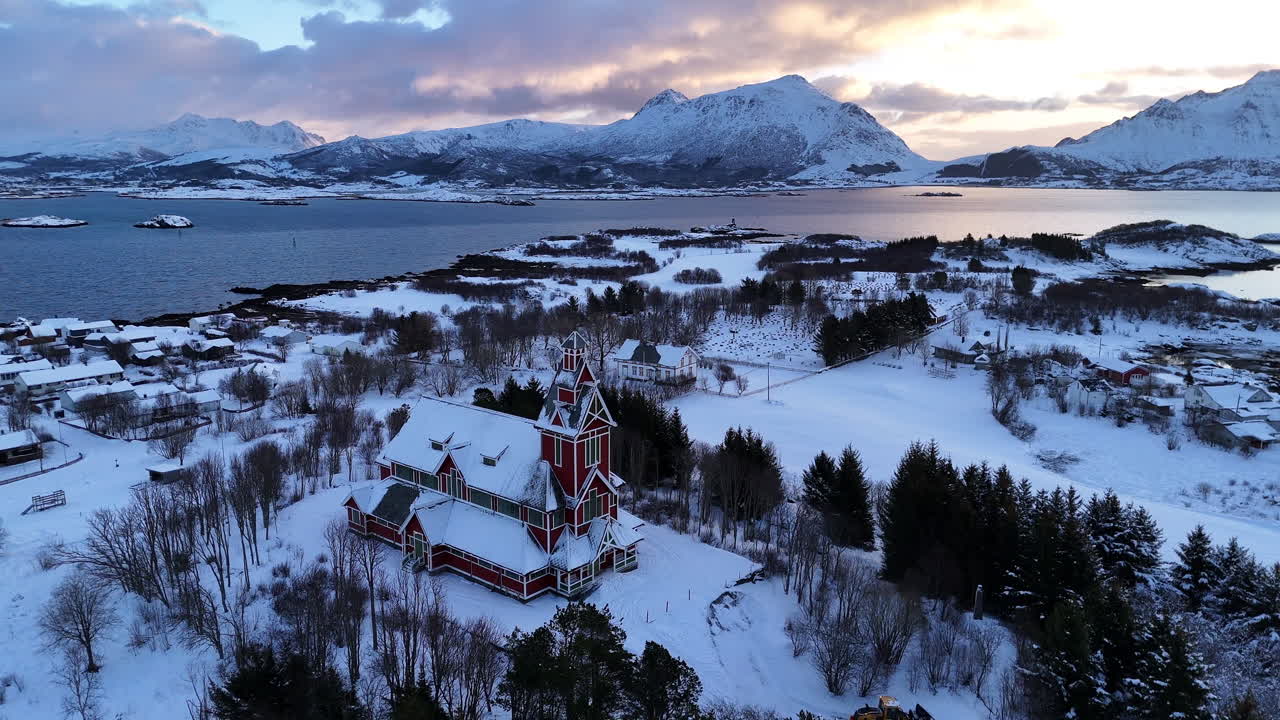 雪に覆われた風景に囲まれた、ノルウェー、ロフォーテン諸島のブクスネス教会の映画のような空撮。赤い木造建築、フィヨルドの海岸線、夕暮れ時のドラマチックな冬の山々。