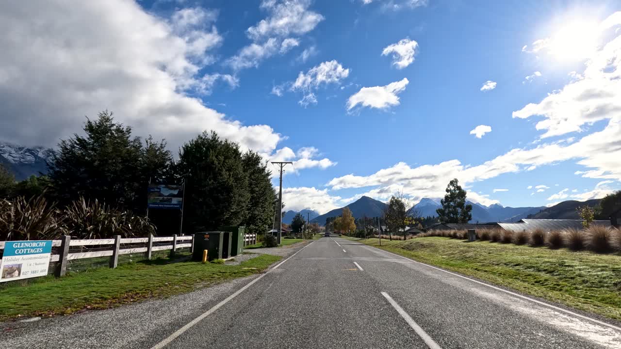 Vehicle travels down sunlit country road with mountain views, blue sky, and scattered clouds
