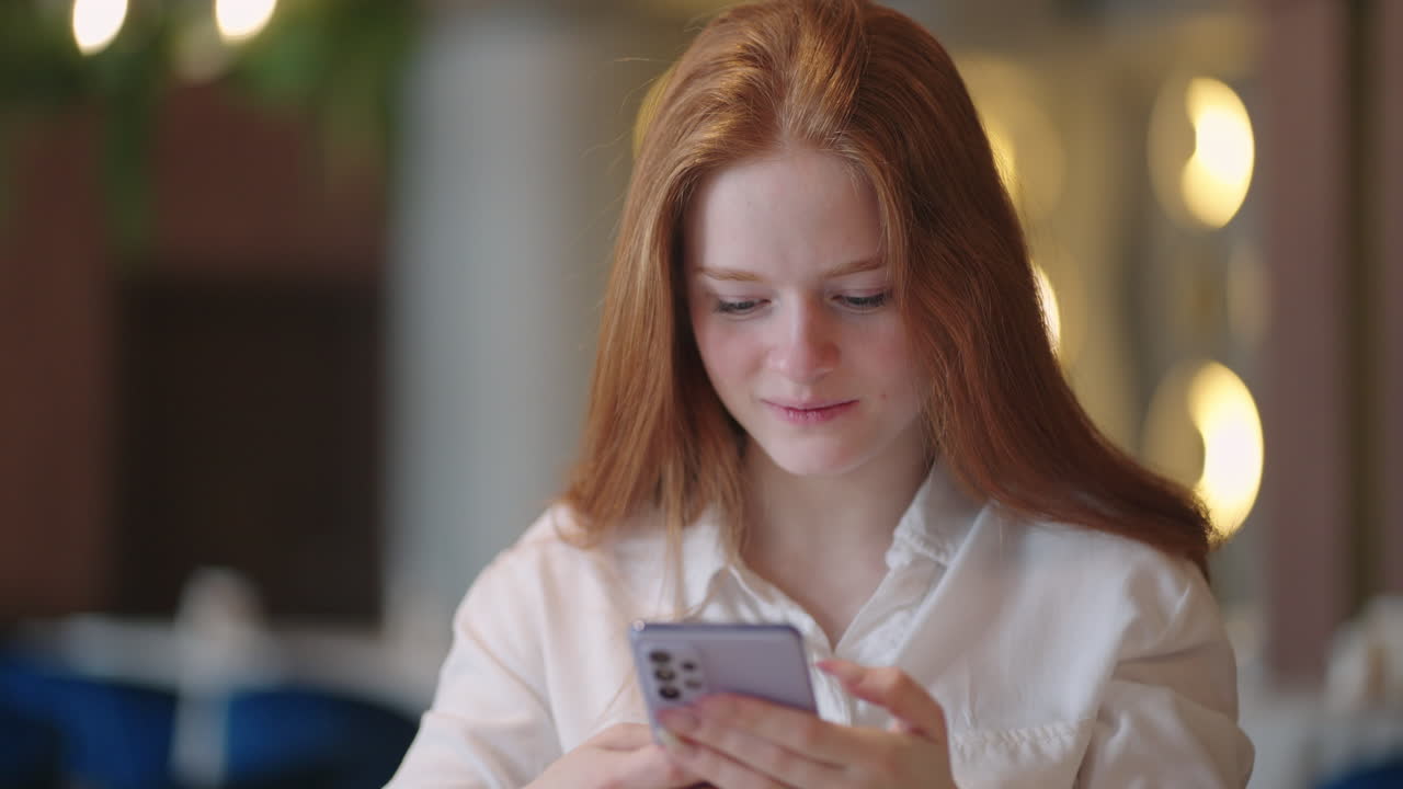 Red-haired Woman working from home using laptop computer while reading text message on mobile phone. Woman using a phone. Serious charming woman using smartphone while working with laptop at home
