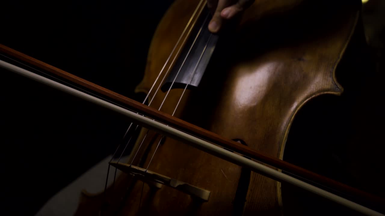 Cellist playing long and legato notes on his cello with black background and overhead light.