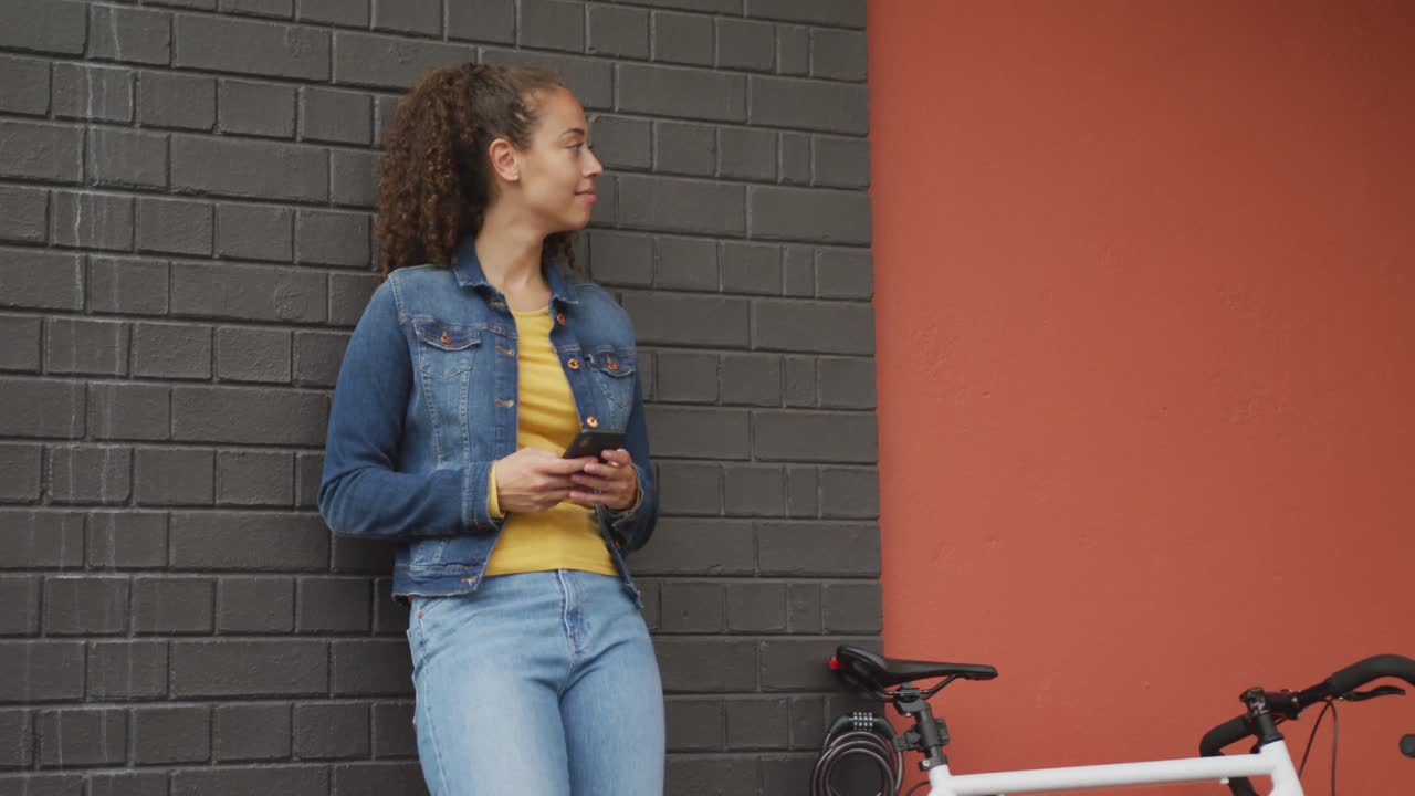feliz mujer biracial en la ciudad, al lado de la bicicleta usando un teléfono inteligente