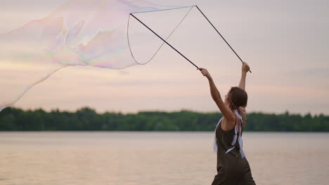 a young female artist shows a soap bubble show blowing up huge soap bubbles on the shore of a lake at sunset. show a beautiful show of soap bubbles in slow motion