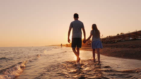 Brother-And-Younger-Sister-Walking-Along-The-Surf-Line-On-The-Beach-At-Sunset