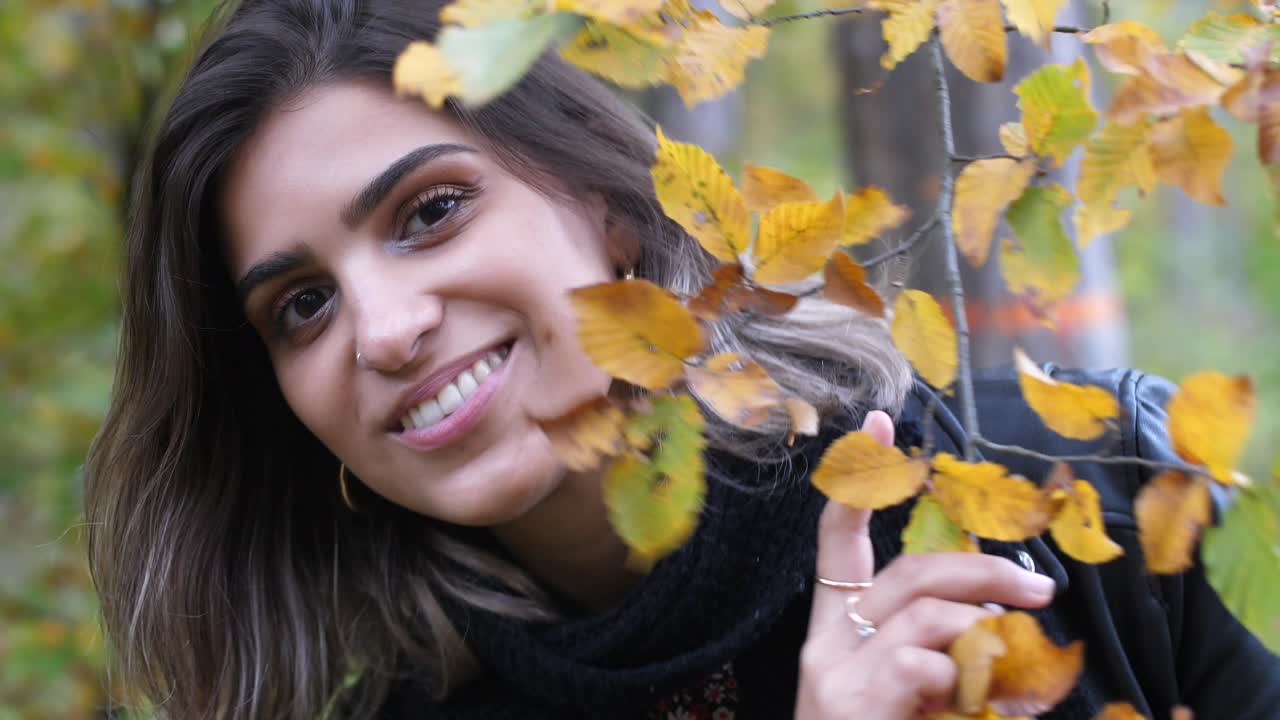 chica morena en el bosque de otoño sostiene una rama con hojas amarillas y se esconde detrás de ella, sonriendo traviesa a la cámara