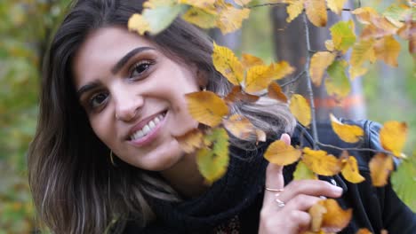 brunette girl in autumn forest is holding a branch with yellow leaves and hiding behind it, smiling naughty at camera