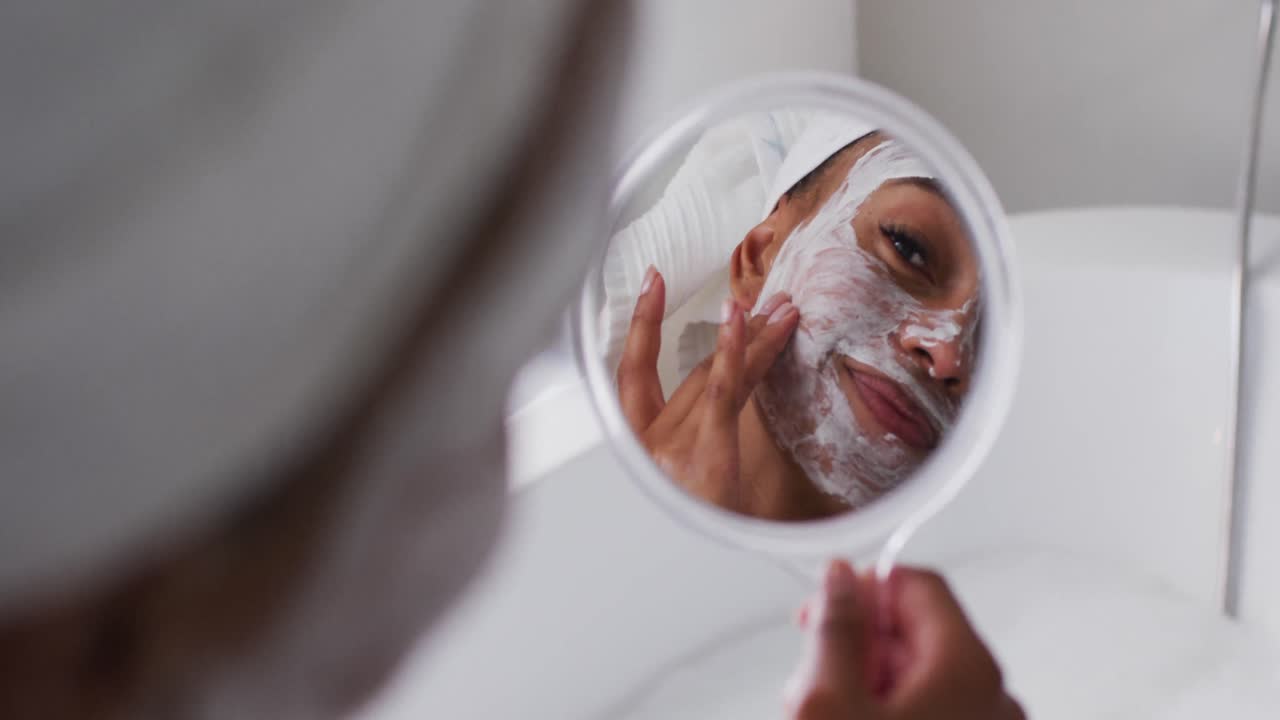 mujer afroamericana aplicando una máscara facial mientras sostiene un espejo en el baño en casa