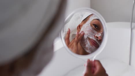 Mujer-Afroamericana-Aplicando-Mascarilla-Mientras-Sostiene-Un-Espejo-En-El-Baño-De-Casa